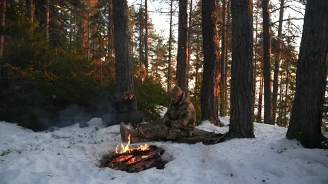 The hunter sits by the fire in the winter forest and drinks from a mug Stockbeeldmateriaal 235427809