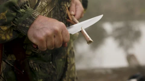 Hunter with a weapon with a sharp knife sharpens a stick shavings from a stick Stock Footage 196623686