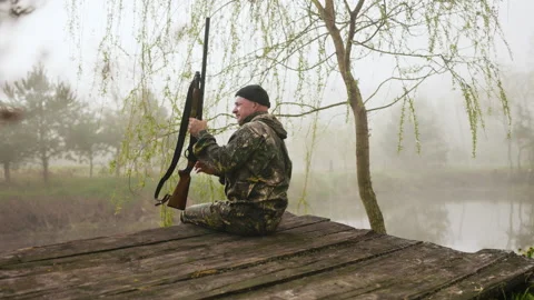 Hunter with a weapon sitting near a tree on a wooden bridge puts his weapon on Stock Footage 196623776