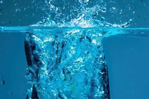 Ice cube dropping into water creating splash and bubbles Stock Photos