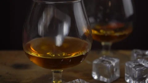 Ice cube falling into cognac \ whiskey glass, golden color strong spirit alcohol Stock Footage 135583406