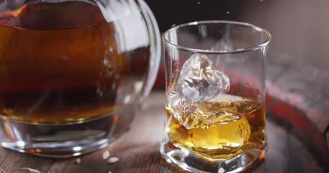 Ice cube slowly falls into a glass of whisky on top of a vintage whiskey barrel. Stock Footage
