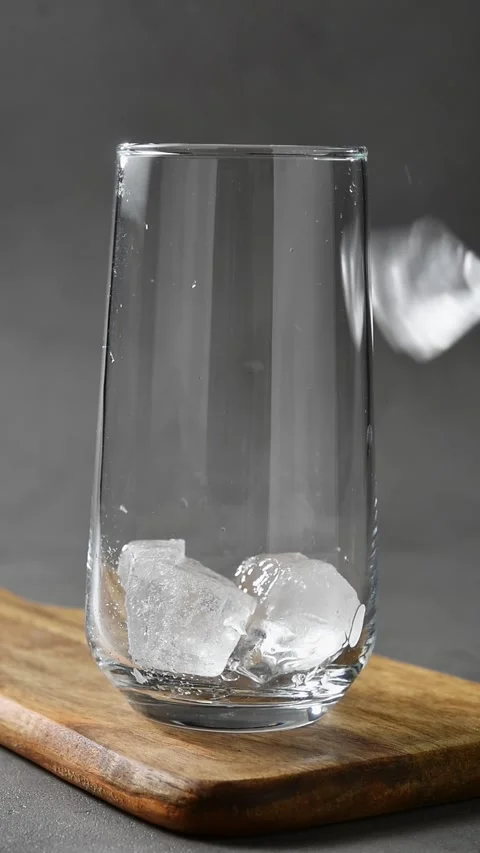 Ice cubes drop into clear glass on wooden board to prepare chilled base for Stock Footage 345486853