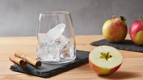 Ice cubes drop into empty glass beside apples and cinnamon before apple cider Stock Footage 345557842