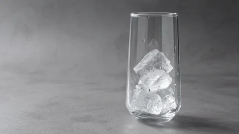 Ice cubes drop into tall clear glass on gray studio background, preparing Stock Footage 345486919