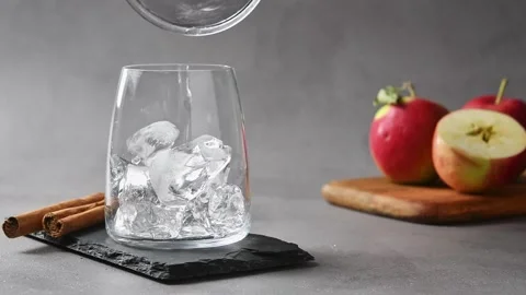 Ice cubes fall into glass on slate board beside apples and cinnamon for cold Stock Footage 345557244
