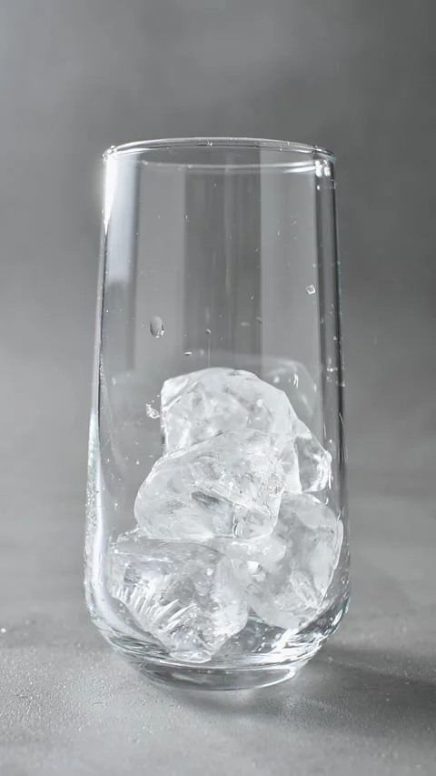 Ice cubes fall one by one into clear drinking glass on gray background in Stock Footage 345486656