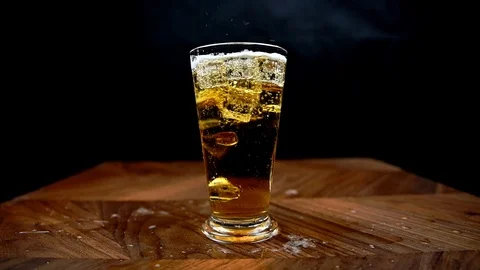 Ice falling into glass of beer in slow motion with beer splashing onto table Video stock 125767609