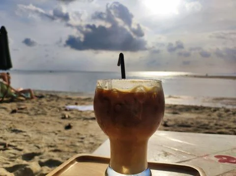 Iced coffee sits on a table at the beach during sunset with people in the b.. Fotos de archivo