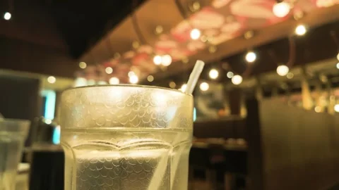 Iced Drink Glass with Straw in Soft Bokeh Restaurant Interior Stock Footage 345370197