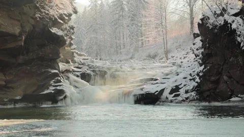 Icy winter waterfall steaming between stones, orange sunlight, frozen pond Stock Footage 100989395