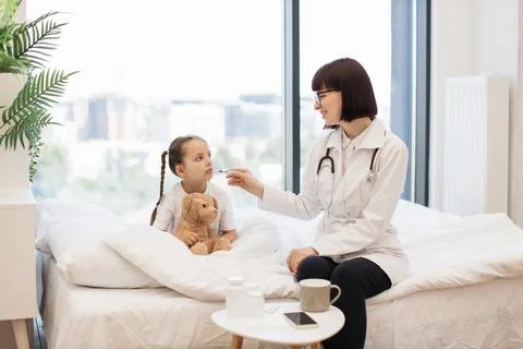 Ill girl resting on bed while doctor measuring temperature Stock Photos