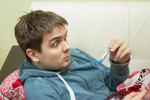 Ill man checking temperature, lying on sofa Stock Photos