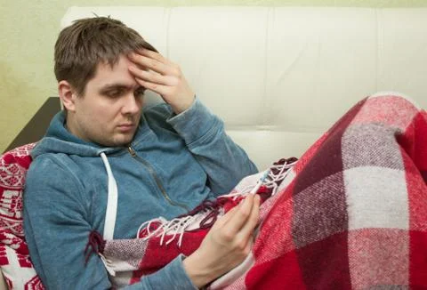 Ill man checking temperature, lying on sofa Stock Photos