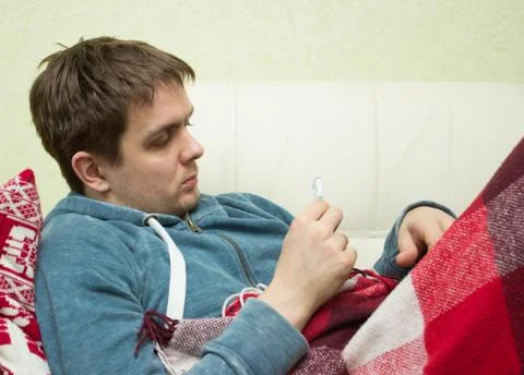 Ill man checking temperature, lying on sofa Stock Photos