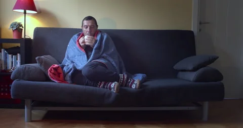 Ill man with flu sitting on the sofa wrapped in a blanket while drinking hot tea Stock Footage 142423672