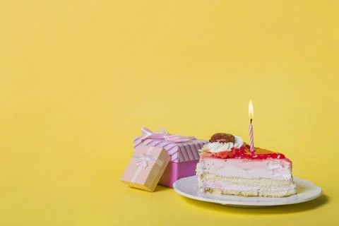 Illuminated candle slice cake with two gift boxes against yellow background Foto stock