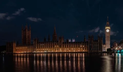 Illuminated clock tower and long gothic building stand beside river under d.. 库存照片