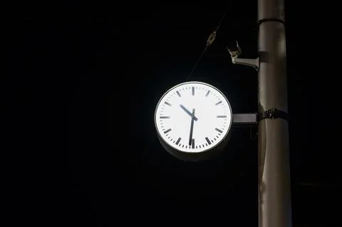 Illuminated public clock face mounted on a pole with a security camera at nig 스톡 사진
