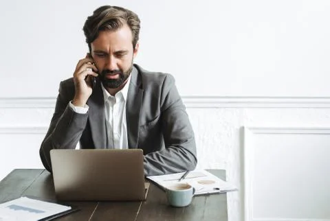 Image of brooding concentrated businessman using laptop and talking on cellph Stock Photos