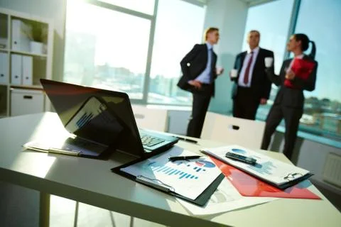 Image of business documents on workplace with three partners interacting on back Stock Photos