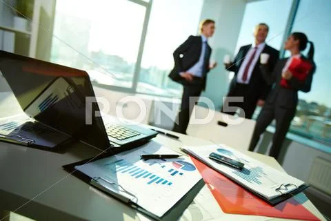 Image of business documents on workplace with three partners interacting on back Stock Photos