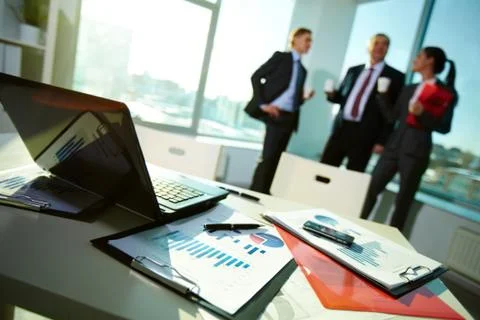 Image of business documents on workplace with three partners interacting on back Stock Photos