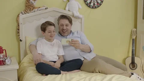 Image of cheerful father using smartphone and lying in bed with little son. Stock Footage 198375517