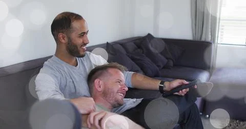 Image of light spots over diverse male couple embracing and watching tv Ilustração Stock