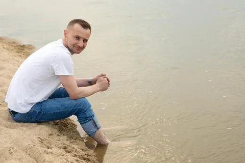 The image lonely positive and smiling man sitting on a beach by the river Stock Photos