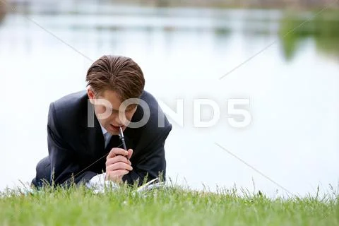 Image of pensive businessman with pen planning work while lying on green grass Stock Photos