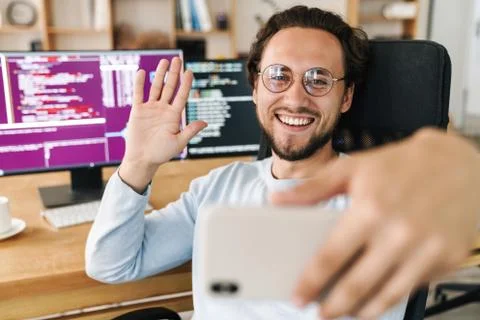 Image of programmer man waving hand while taking selfie on cellphone Stock Photos