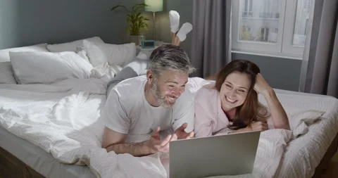 Image of romantic cheerful couple using laptop and chatting while lying on bed Stock Footage 156460093