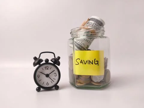 The image shows a glass jar filled with coins and alarm clock Stock Photos