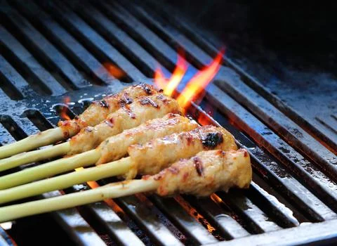 The image shows a row of five chicken satay skewers being grilled on a grill. Stock Photos