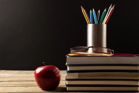 Image of stack of books, crayons in container and apple on black background Stock Photos