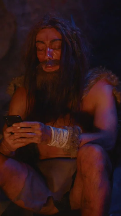 Imagine a prehistoric man humorously using a modern smartphone inside a cave Stock Footage 310814923