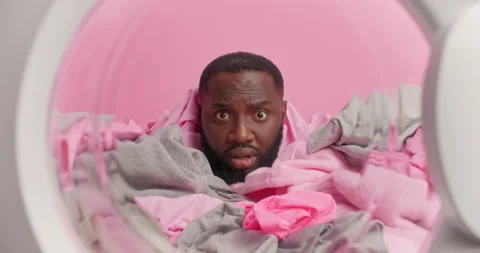Impressed dark skinned bearded man sticks head from pile of laundry does washing Stock Footage 156632180