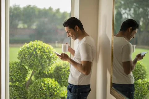 India, Man at window using mobile phone and holding coffee cup Stock Photos