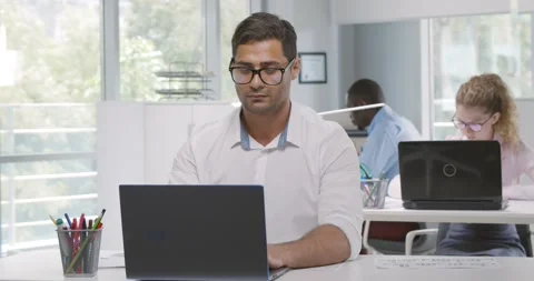 Indian financier sitting at desk using laptop in busy corporate office Stock Footage 157866865