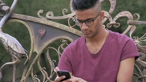 Indian Guy in Glasses Writes a Message on a Smartphone Sitting on a bench in the Stock Footage 95413935