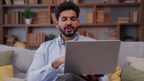 Indian guy sit on sofa at home look at camera wave hand greets friend make vi Video stock 238541318