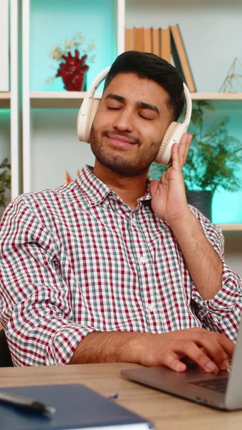Indian man on home table typing on laptop with headphones listening to music Stock Footage 347055735
