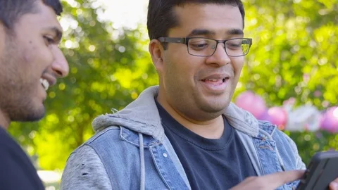 Indian man laughs at something on his phone with his friend at the park - Slow Stock Footage 125090282