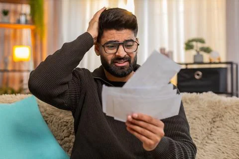Indian man looking through receipts with worry on sofa at home, financial stress Stock Photos