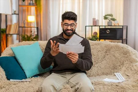 Indian man looking through receipts with worry on sofa at home, financial stress Stock Photos