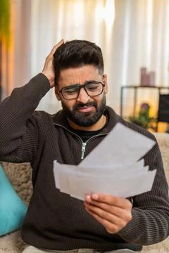 Indian man looking through receipts with worry on sofa at home, financial stress Stock Photos