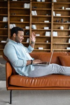 Indian man is lying down on the sofa and using trendy laptop for video call, Stock Photos