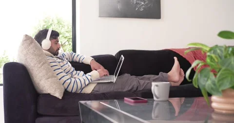 Indian man reclining on dark sofa working on laptop while listening through Stock Footage 338325939