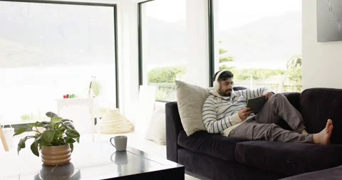 Indian man settling on sofa by windows, putting on headphones, tapping tablet to Stock Footage 344948237